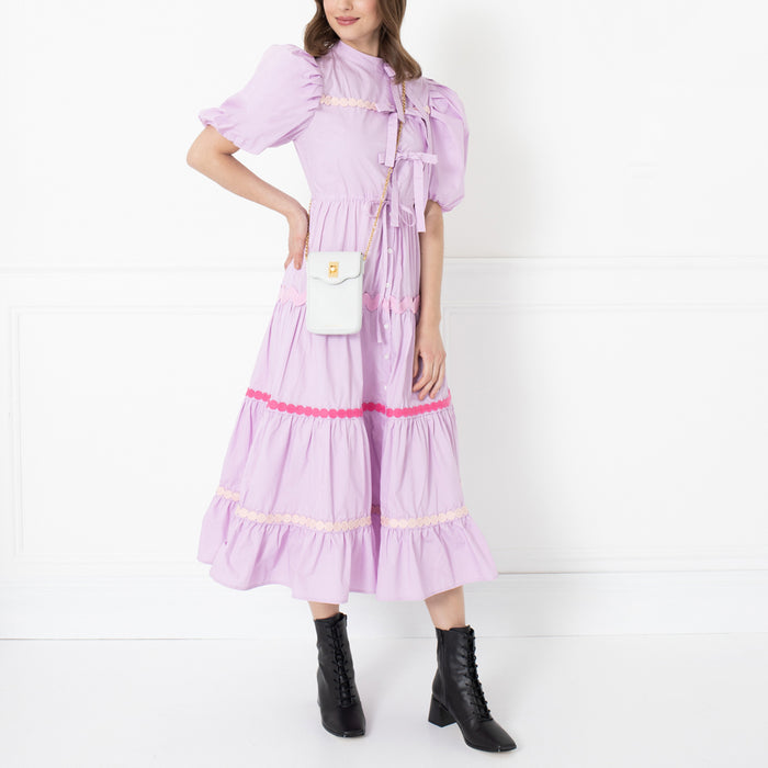 A woman in a lavender tiered midi dress with pink and cream detailing, carrying a white crossbody bag and wearing black ankle boots, against a white background.