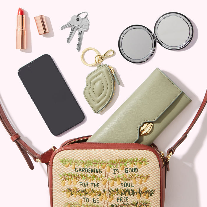 A beige crossbody bag with embroidered pears and the text "Gardening is good for the soul to be free" lies open on a white background, revealing a sage green wallet, a smartphone, keys, a compact mirror, and lipstick.
