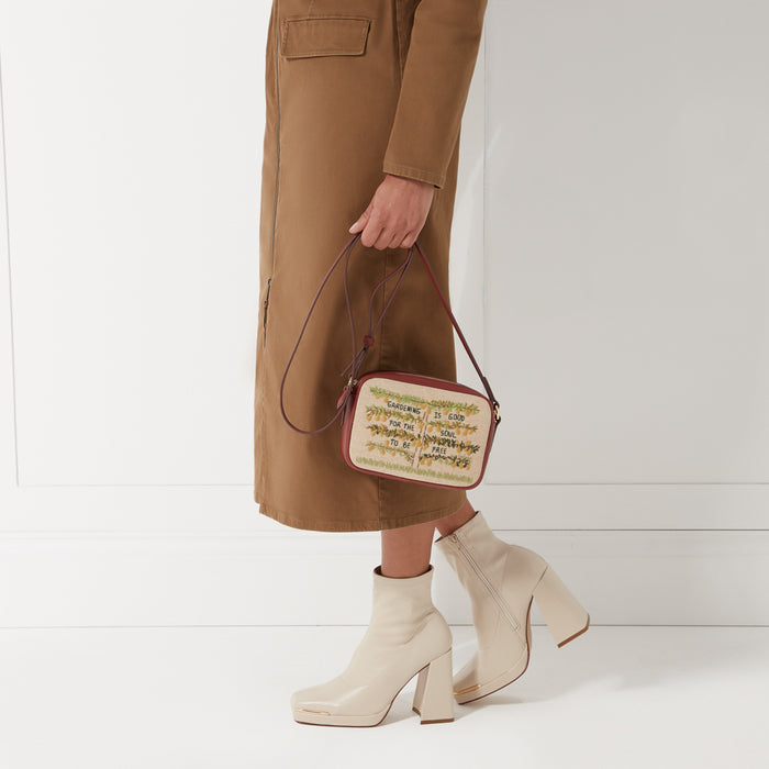 Woman in brown midi dress holding a rectangular crossbody bag with embroidered text that says "Gardening is good for the soul to be free". She's wearing cream-colored boots with thick block heels. The background is white.