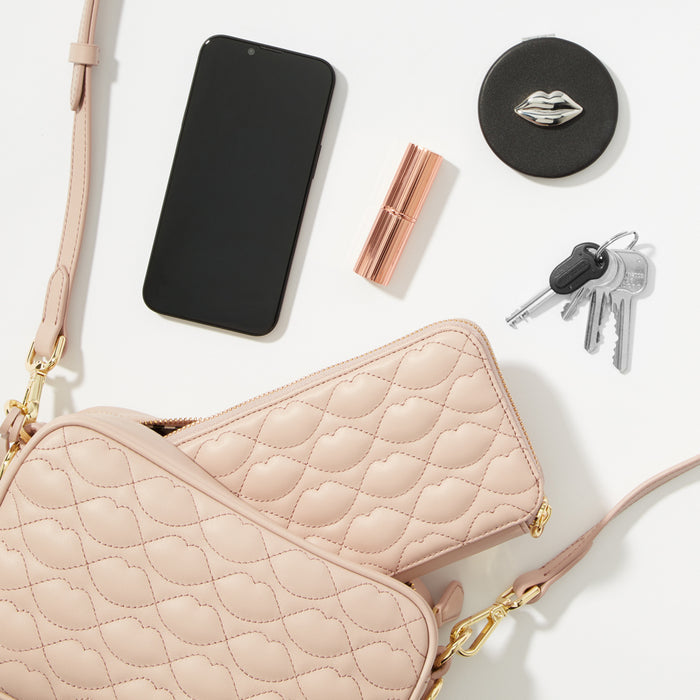 A blush pink quilted crossbody bag with a matching wallet inside, alongside a smartphone, lipstick, compact mirror, and keys, all arranged on a white background.