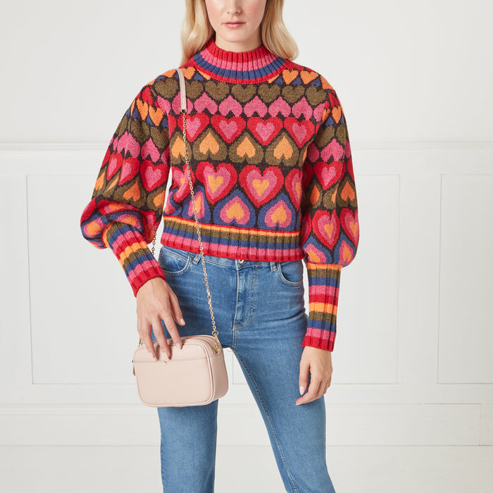 A woman in a multi-coloured heart-patterned sweater and light blue jeans carries a pale pink crossbody bag.