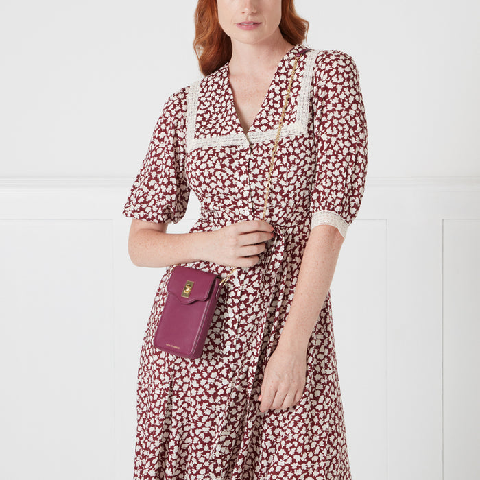 A woman in a floral dress holds a dark burgundy crossbody phone bag with gold hardware. The background is white.