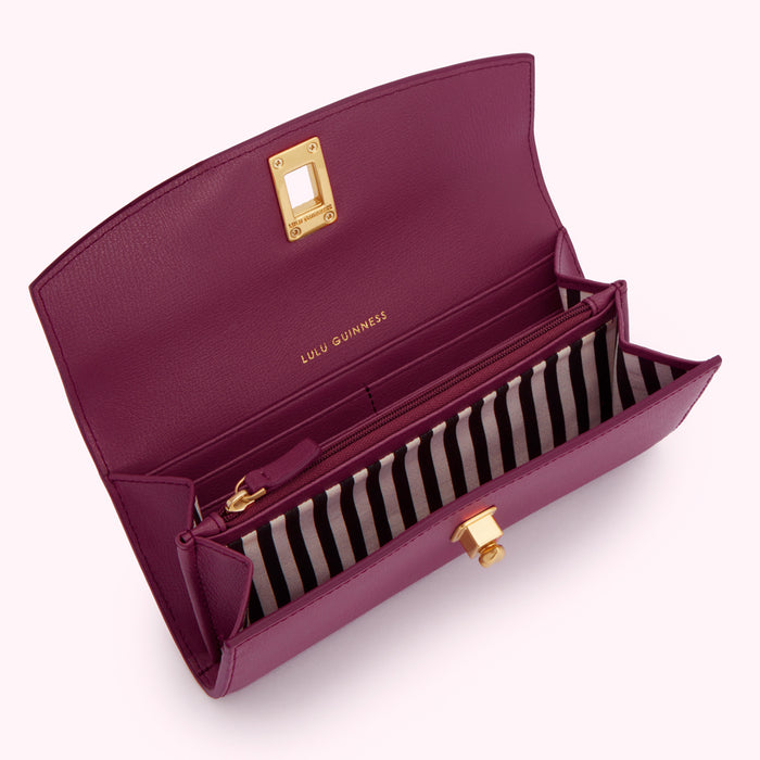 Open plum-coloured designer wallet with gold-tone hardware, displayed against a light pink background, showing an interior lined with black and white striped fabric, multiple compartments, and a zippered pocket.