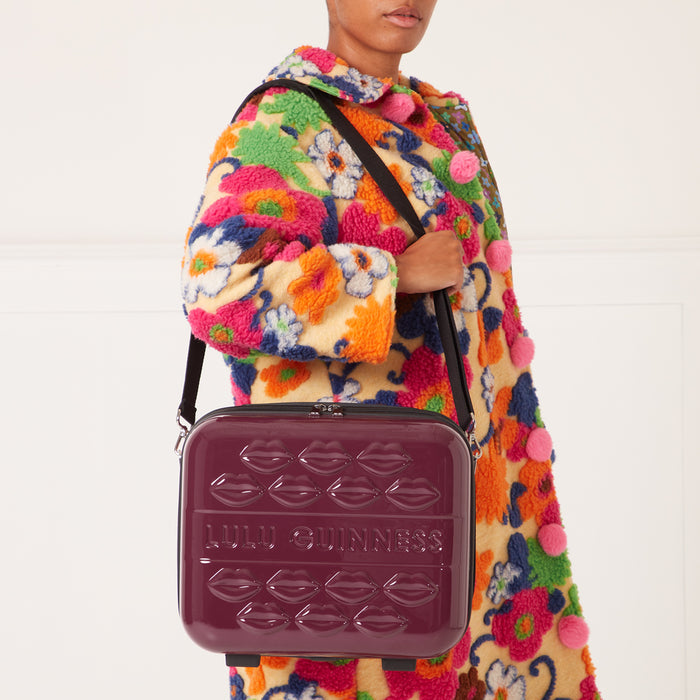 A burgundy Lulu Guinness makeup case with a lip design, carried by a model wearing a colourful floral coat.