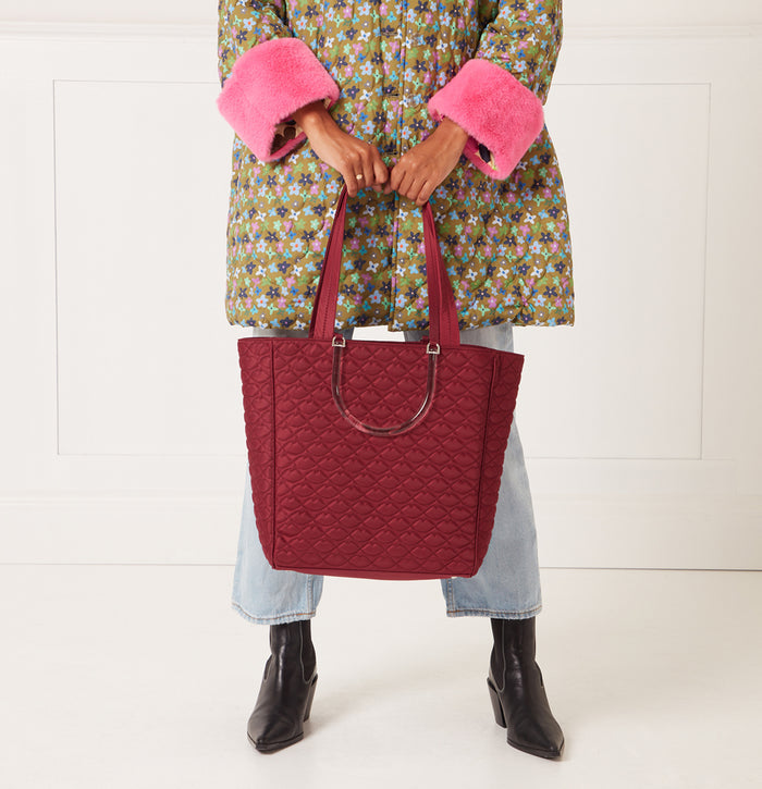 A woman in a floral quilted coat and light blue jeans holds a large burgundy quilted tote bag with translucent handles.