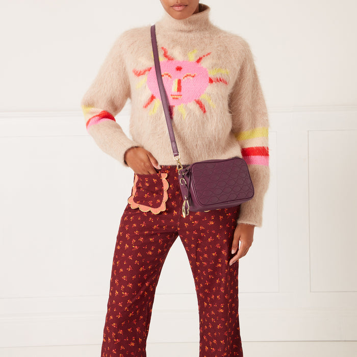 A woman models a deep purple quilted crossbody bag against a white background. She is wearing a fluffy beige sweater with a pink sun design and burgundy floral pants.