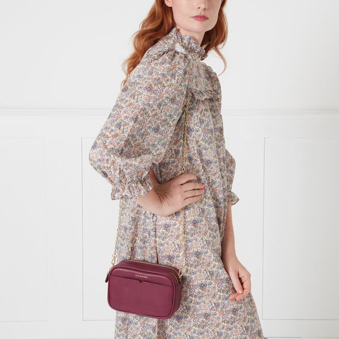 A woman in a floral dress carries a burgundy crossbody bag with a gold chain strap against a white background.