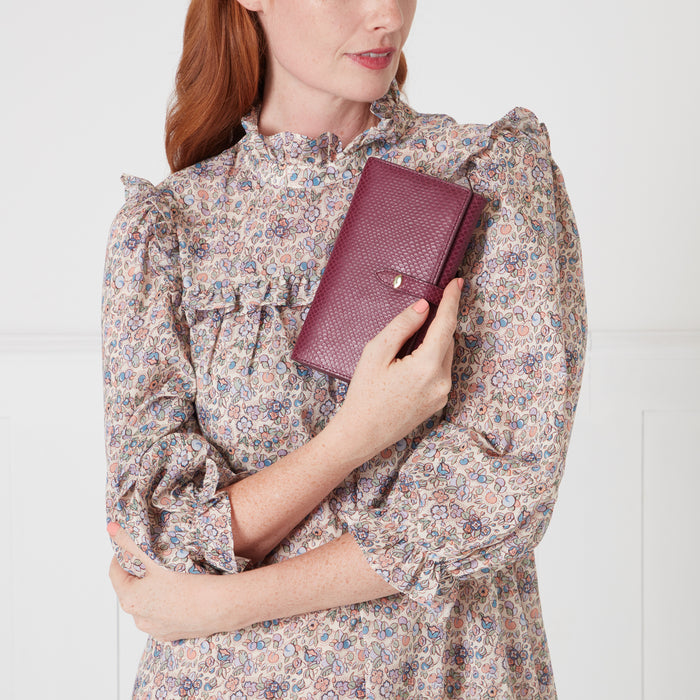 Woman with auburn hair wearing a floral dress holds a burgundy leather wallet. The background is off-white.