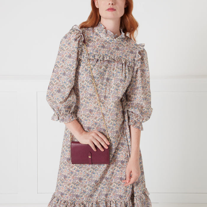 A woman with red hair models a burgundy leather wallet-on-chain against a white background while wearing a floral dress.