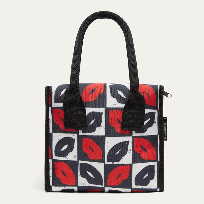 A square lunch bag with black handles and a pattern of red and black lips on a white and dark gray checkered background, shown against a light gray studio backdrop.