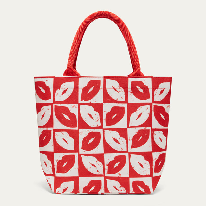A red and white tote bag featuring a repeating pattern of lips printed in red on a white background, with red fabric handles, shown against a light gray studio background.