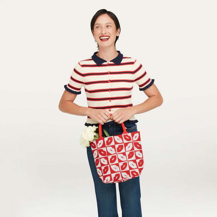 A woman wearing a red, navy, and cream striped polo shirt and blue jeans holds a red and white patterned tote bag with red handles and a checkerboard lip print. She is standing against a plain light gray studio background.