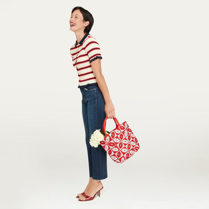 A woman wearing a red, white, and navy striped short-sleeve polo shirt and blue jeans holds a red and white checkerboard lip patterned tote bag with white flowers tucked inside, posed against a solid off-white studio background.