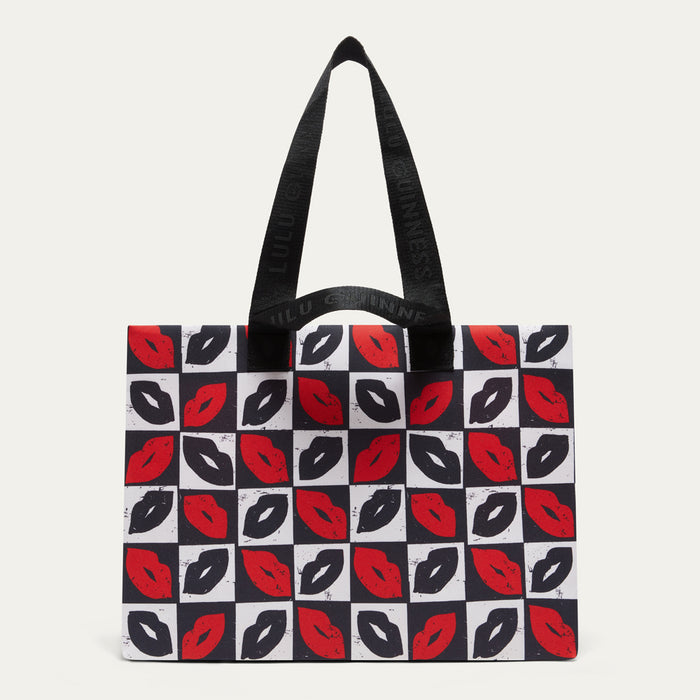 A tote bag featuring a black, white, and red lips pattern on a checkerboard design, with black shoulder straps, photographed against a light gray background.