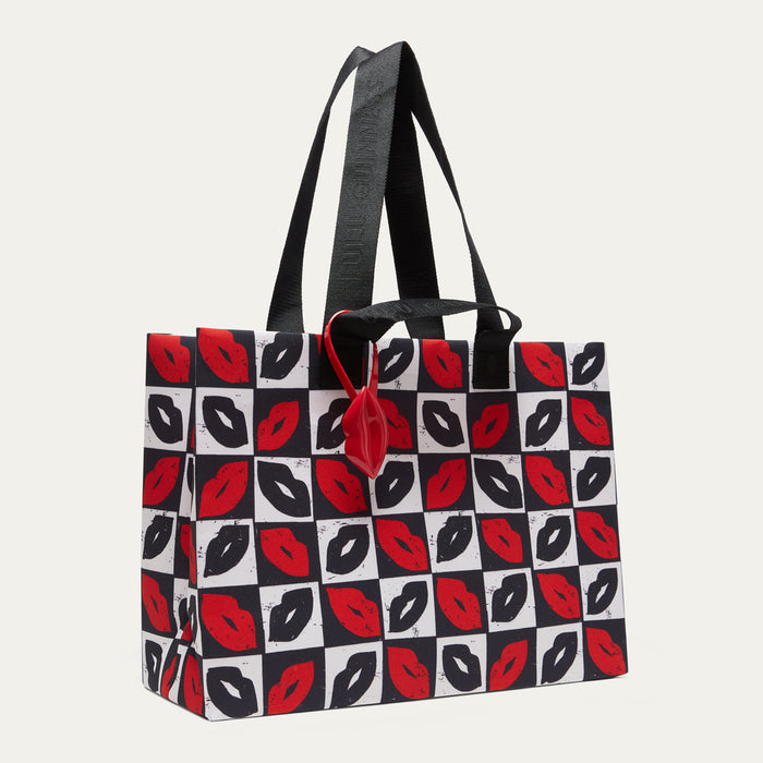 A tote bag with a black and white checkerboard pattern featuring red lips print, black shoulder straps, and a red hanging tag, shown against a light gray background.