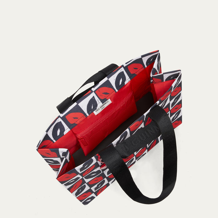 A tote bag with a bold black, white, and red lip print exterior, a solid red interior, and black webbed handles, shown on a light off-white background.