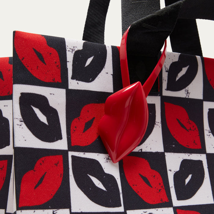A close-up of a tote bag featuring a black, white, and red lip print pattern, with a glossy red lip-shaped charm and black handles.