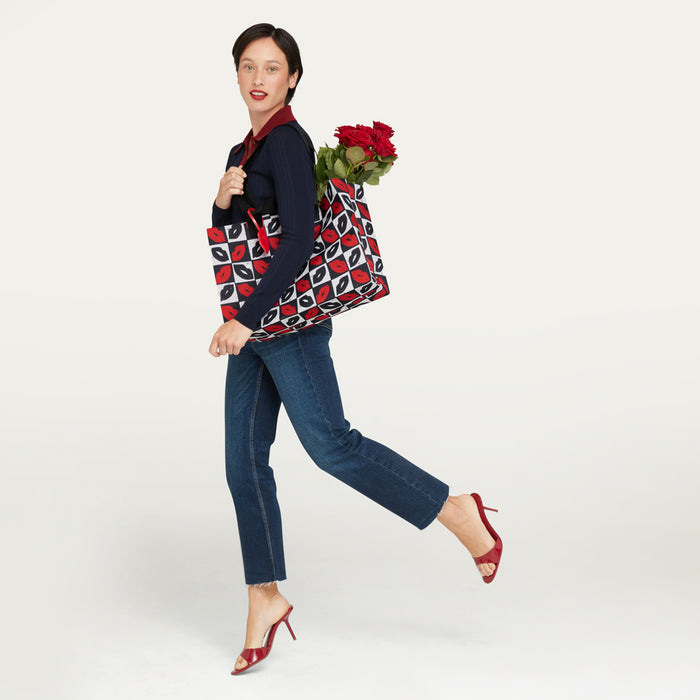 A woman in a navy sweater, blue jeans, and red heels carries a large tote bag with a red, black, and white geometric pattern while holding red roses, photographed against a plain light gray studio background.