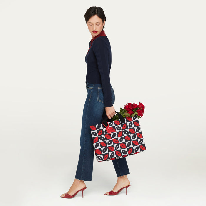 A woman in a navy long-sleeve top, blue jeans, and red heels carries a black tote bag with a red and white geometric lip pattern and red roses, photographed against a clean white studio background.