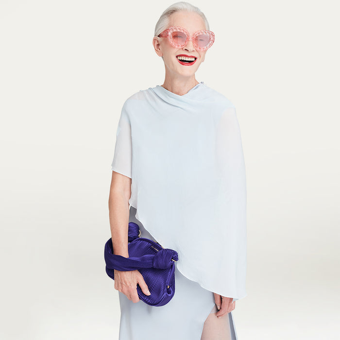 Person wearing a light blue outfit with pink sunglasses and holding a purple satin bag on a light gray background