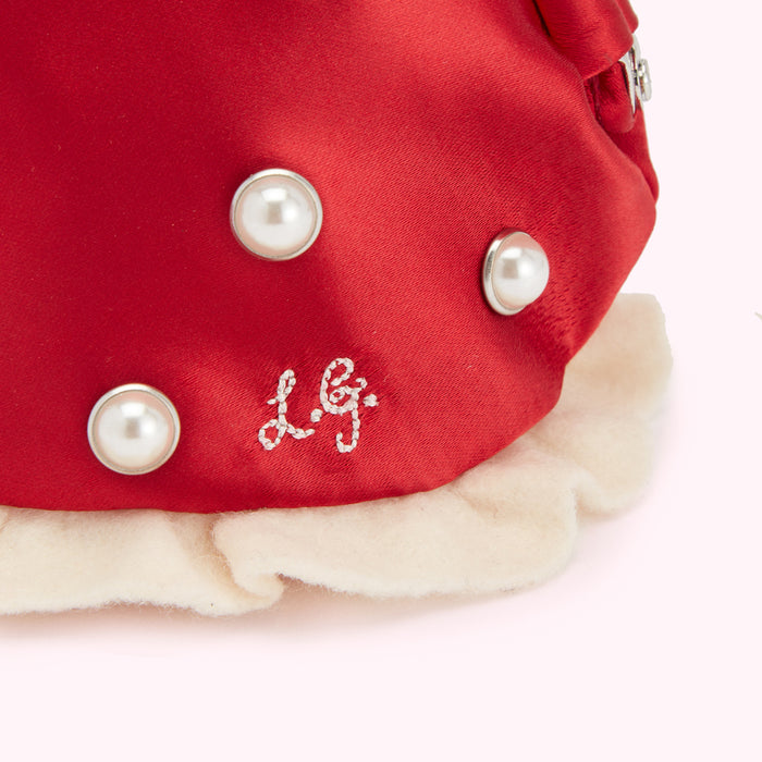 Close-up of a red satin accessory adorned with pearls and the embroidered initials "L.G." on a cream-colored ruffled trim.