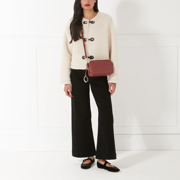 A woman in a cream jacket and black pants wears a maroon quilted crossbody bag and black shoes against a white background.