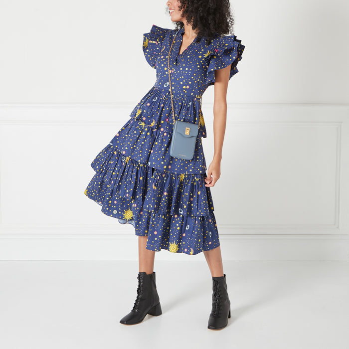 Woman in a blue celestial print dress with ruffled skirt and a blue crossbody bag. She's wearing black lace-up boots. The background is white.