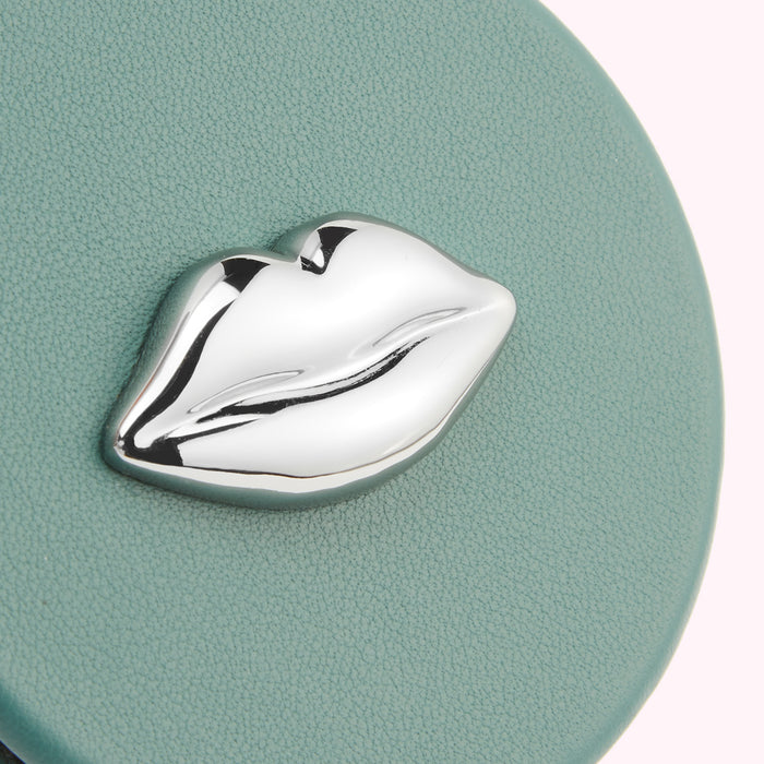 Close-up of a teal circular compact mirror with a silver lip-shaped embellishment.