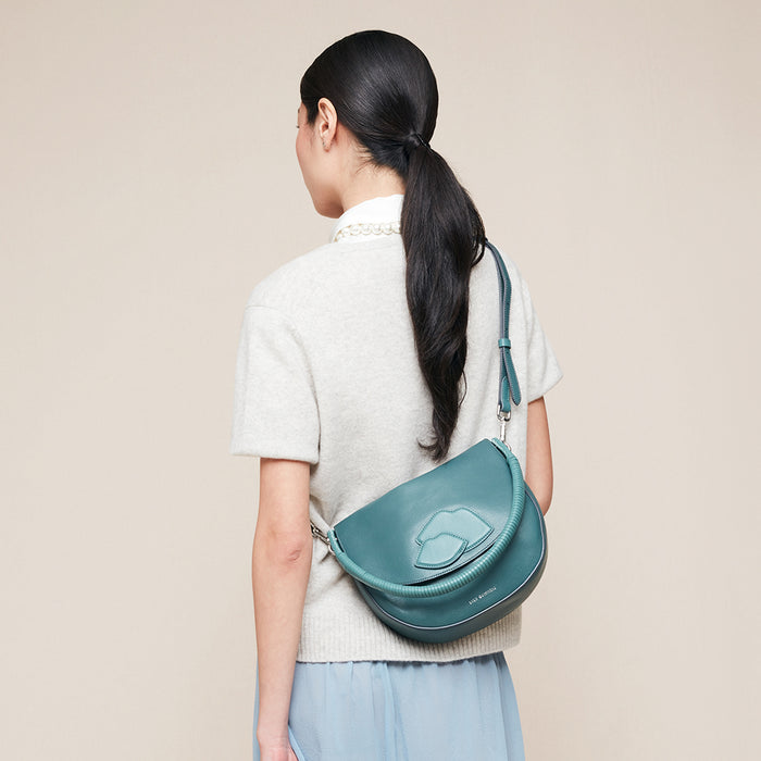 Person wearing a teal leather crossbody bag with a lip design on the front on a beige background