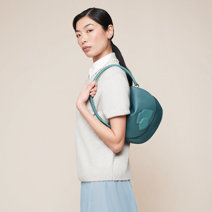 Woman carrying a teal leather shoulder bag with a lip design on the front against a beige background
