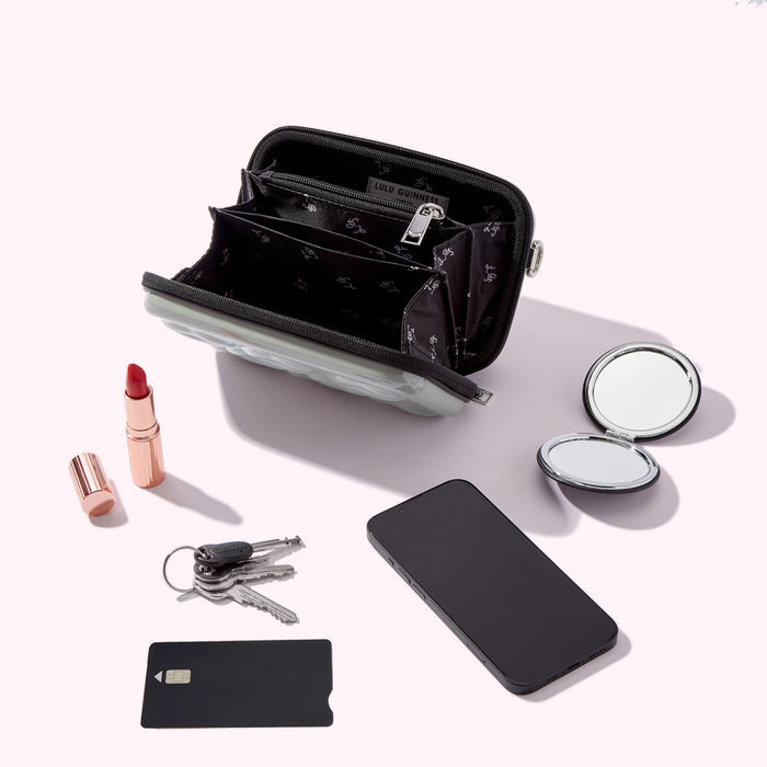 A pale grey Lulu Guinness zipped purse lies open, revealing its black interior compartments. Beside it are a lipstick, keys, a compact mirror, and a smartphone; a black credit card lies in the foreground. The background is a solid, light colour.