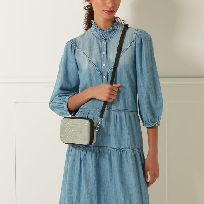 A light sage green crossbody bag with a black strap and embossed lip pattern is shown on a model wearing a light wash denim dress.