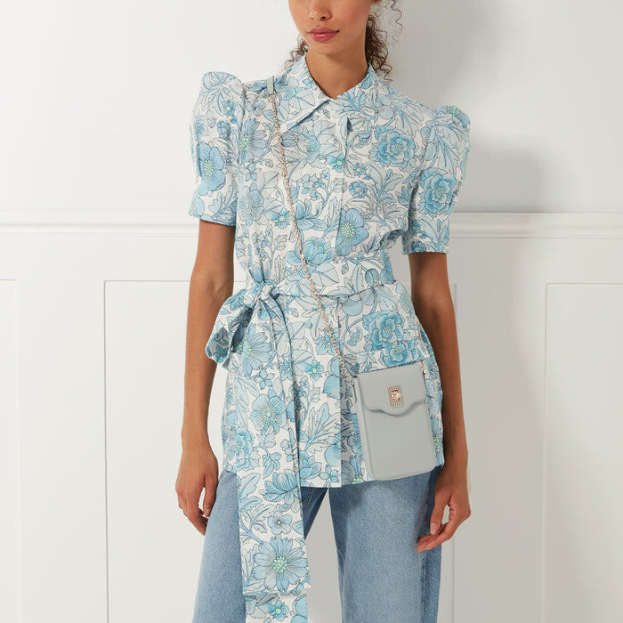 A woman wearing a light blue floral shirt dress and light blue jeans, with a light blue crossbody phone bag.