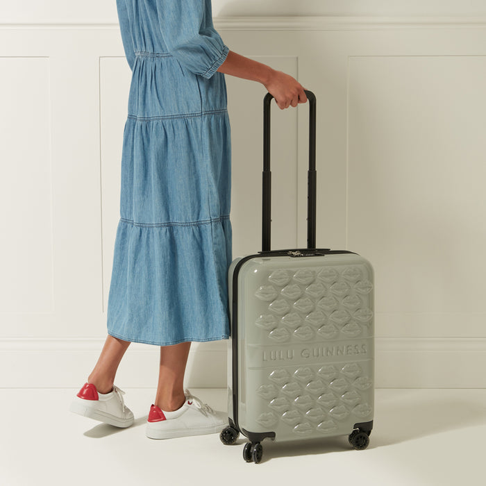A light grey Lulu Guinness carry-on suitcase with a lip print design, being held by a person wearing a denim dress and white sneakers with red accents against a white background.