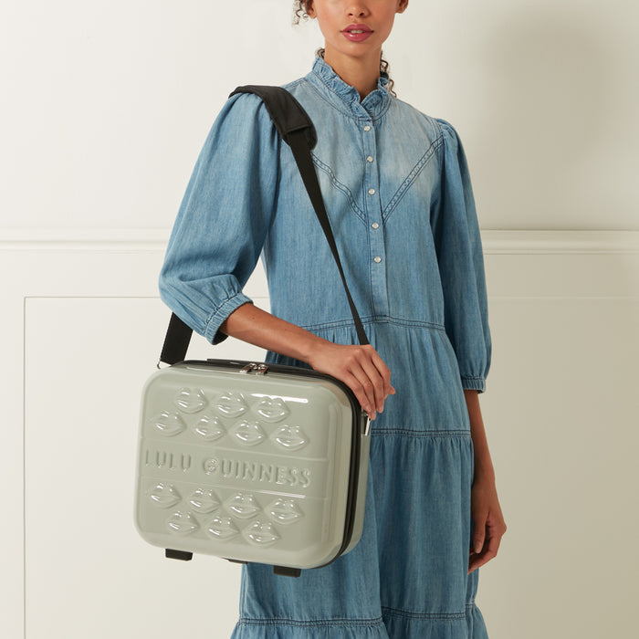 A woman in a denim dress holds a light grey Lulu Guinness makeup case with a black shoulder strap and a lip design.
