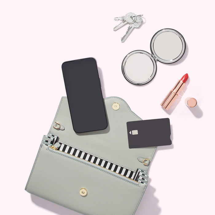 A light grey purse lies open, displaying a smartphone, credit card, and compact mirror. Keys and lipstick are nearby on a light pink background.