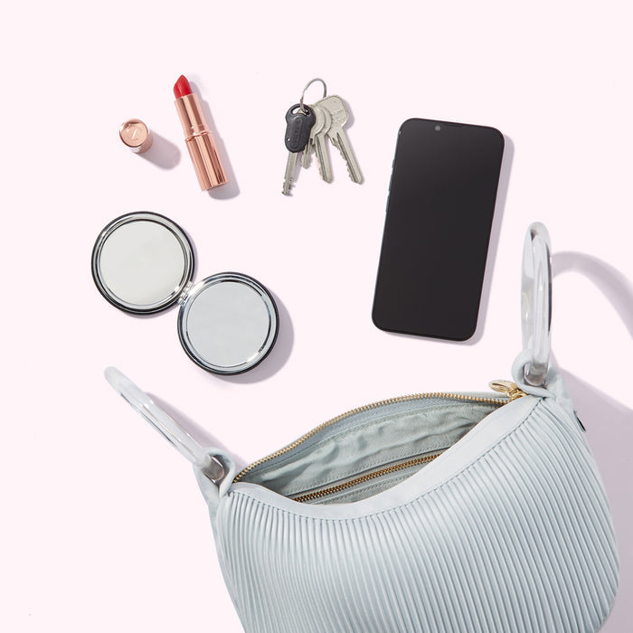 A light blue pleated handbag sits open, displaying its interior. Surrounding it are a compact mirror, lipstick, keys, and a smartphone, all arranged on a white background.