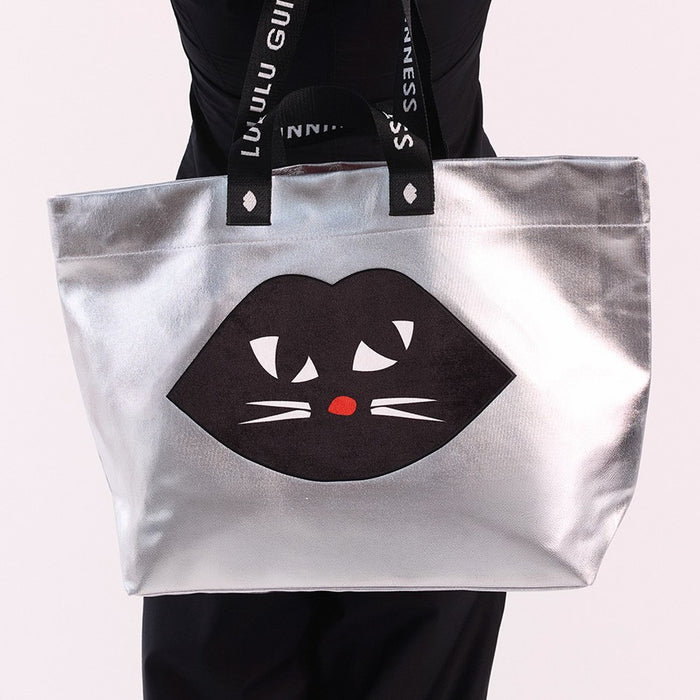 A silver metallic tote bag with black handles featuring the text 'LULU GUINNESS' and a large black cat graphic with white whiskers and a red nose on the front, held by a person wearing a black top and black leggings against a light pink background.