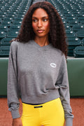 A woman with long curly dark hair stands in a stadium wearing a heather gray crewneck sweatshirt with a small white oval logo on the left chest, paired with bright yellow pants.
|M1