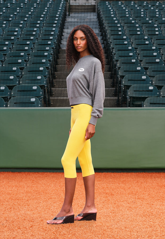 A woman stands on baseball diamond in front of green stadium seating, wearing a gray sweatshirt, bright yellow cropped leggings, and black wedge sandals.
|M1