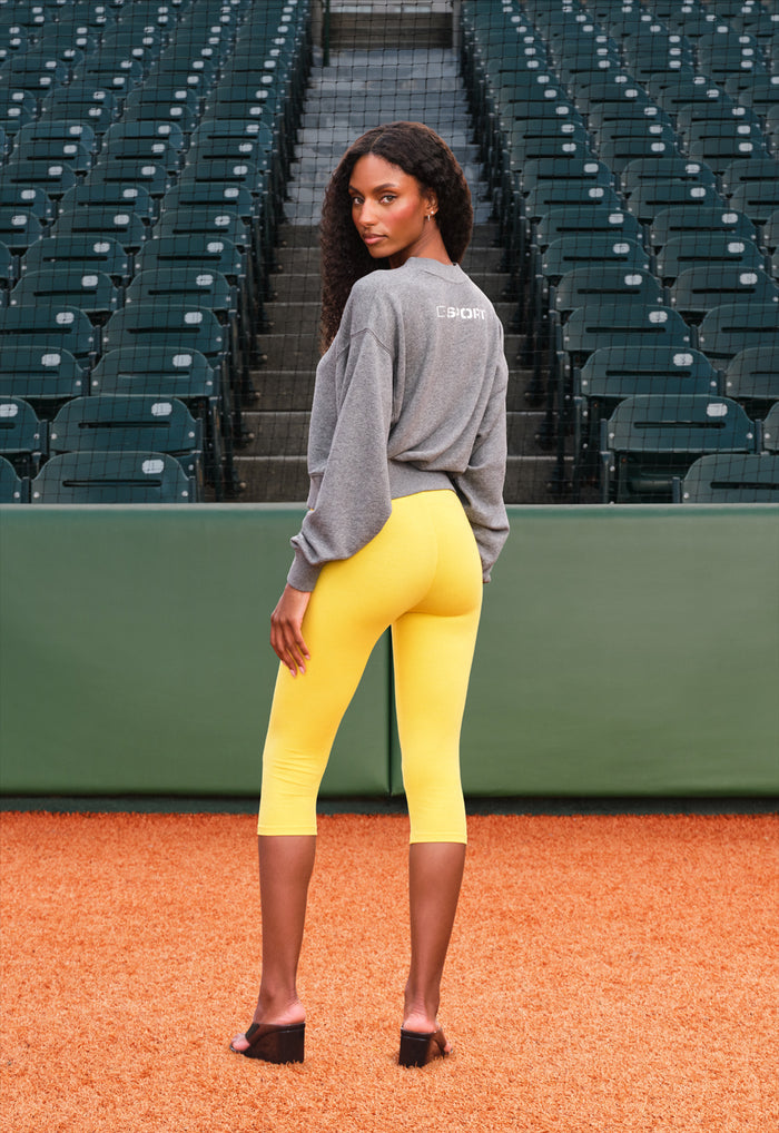 A woman stands on a baseball diamond wearing a gray long-sleeve athletic top and bright yellow fitted capri leggings, photographed from the back. Empty stadium seating and a green wall are in the background.
|M1