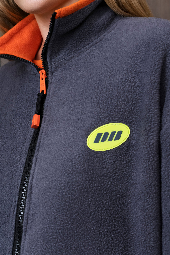 Close-up of a person wearing a dark gray fleece zip jacket with a bright orange collar and zipper pull, featuring a lime green oval logo patch on the chest.