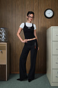 A woman wearing a black ribbed tank top and matching black wide-leg pants stands in an office-like room with wood-paneled walls, a water cooler, and a filing cabinet.
|M1