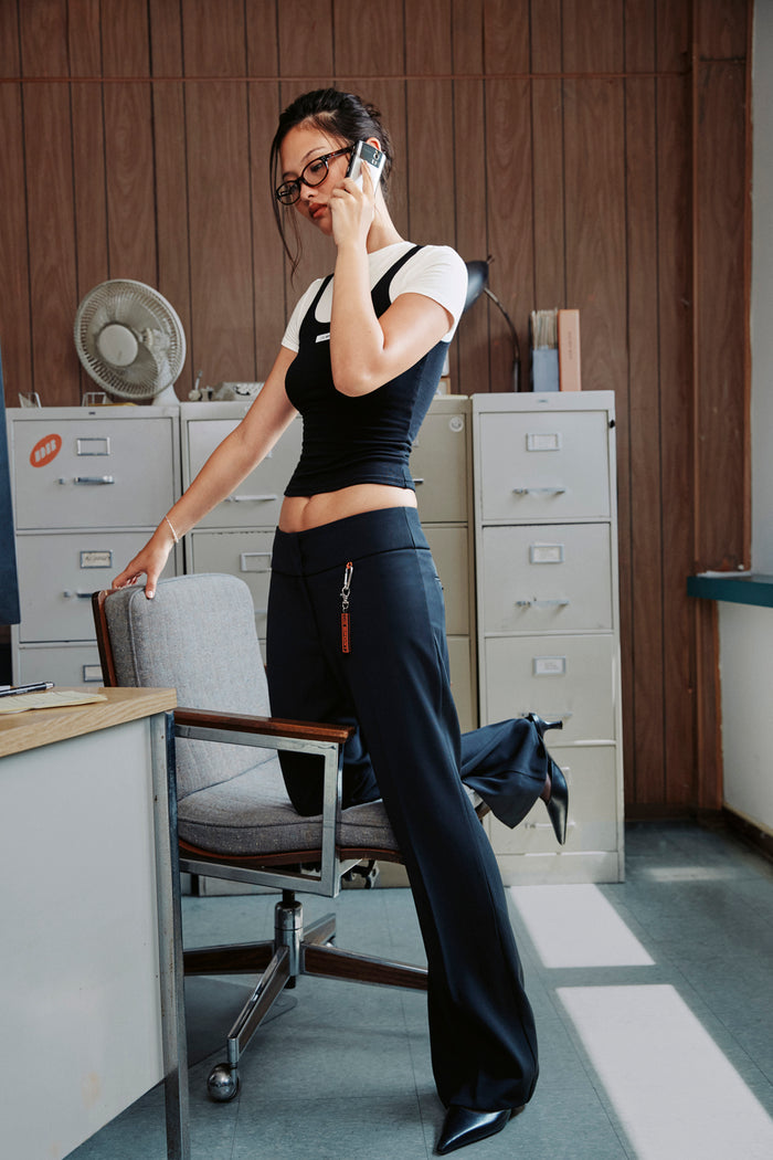 A woman in a black crop top and wide-leg navy pants stands in an office setting, leaning on a chair while talking on a phone.
|M1