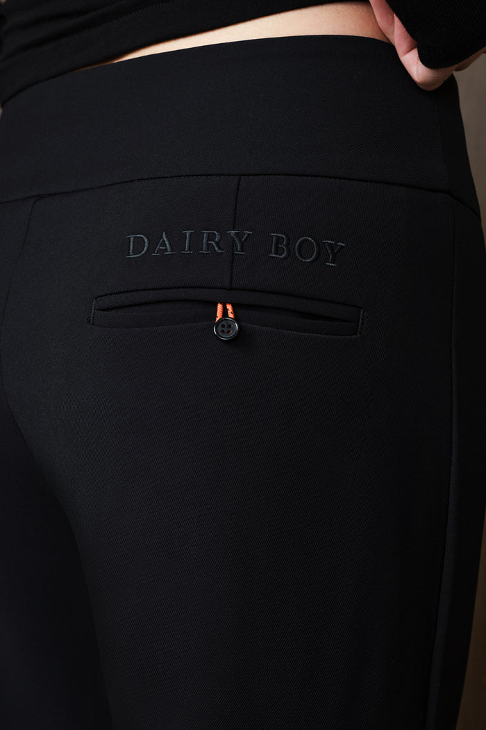 Close-up of black trousers featuring an embroidered 'DAIRY BOY' logo and a zippered pocket with a small orange accent tab, worn by a model.
|M1