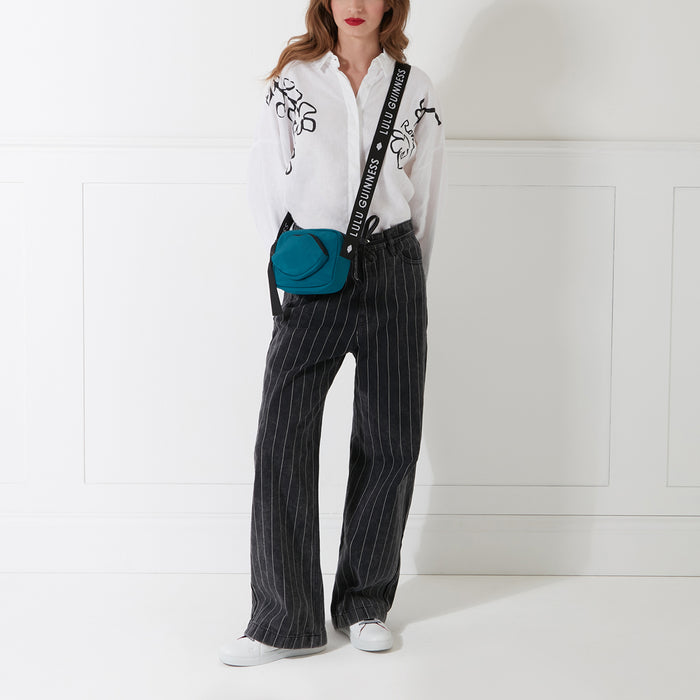Woman wearing a white shirt, black pinstripe pants, white sneakers, and a teal crossbody bag with a black strap that says Lulu Guinness. The background is white.