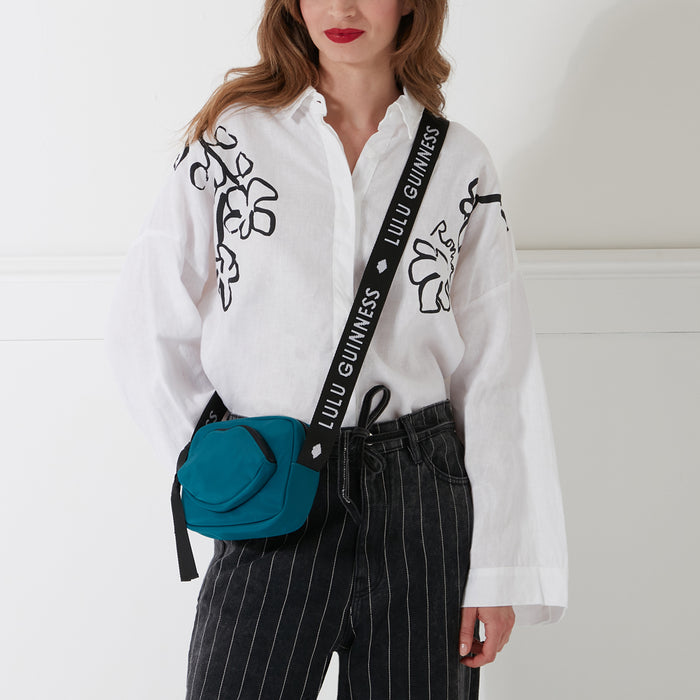Woman with red lipstick wearing a white shirt with black floral details and dark grey pinstripe pants; she is modelling a teal crossbody bag with a black strap that says "Lulu Guinness" on it. The background is white.