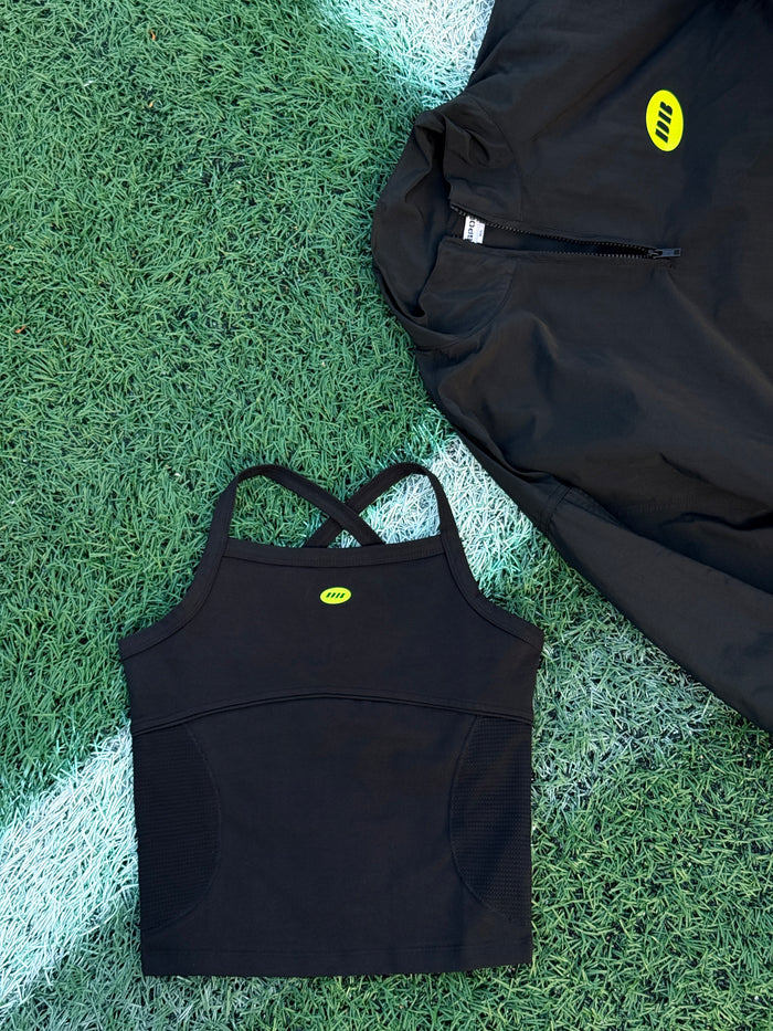 A black athletic zip-up jacket and a matching black sports bra laid flat on green turf with a white line, both featuring a small yellow logo.