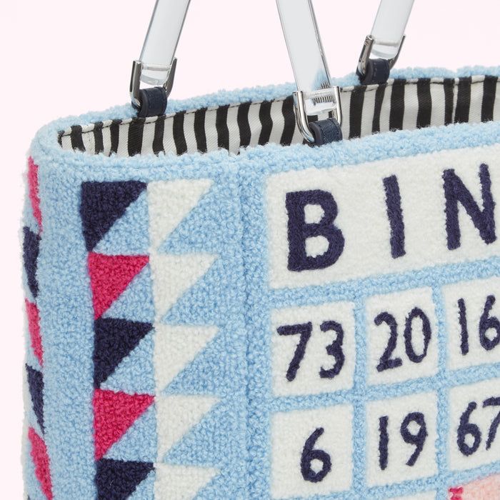 Close-up of a light blue textured handbag with a bingo-themed design, featuring the word "BINGO" and numbers embroidered on it. The bag has clear acrylic handles.