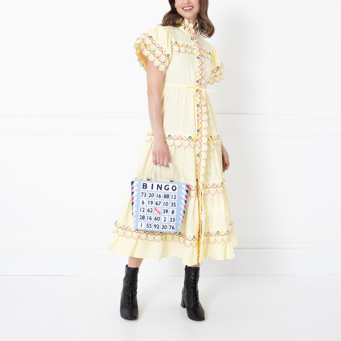 Woman in pale yellow dress holding a bingo card-themed handbag against a white background.